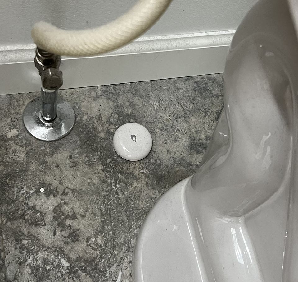 A small round white Aqara water leak sensor on a tile floor behind a toilet, next to the shutoff valve and braided supply line